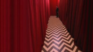 Agent Dale Cooper (played by actor Kyle Mcklachlan) at the end of a long corridor of red curtains with a black and white zig zagged floor in the black lodge in twin peaks